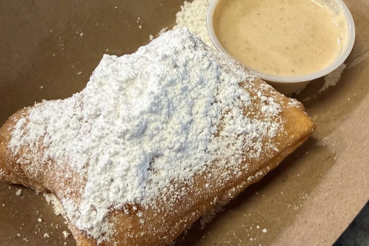 Powdered sugar beignet with dipping sauce in a cardboard tray.