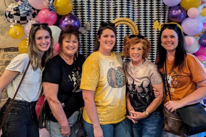 Five women smiling in front of a balloon arch and geometric backdrop.