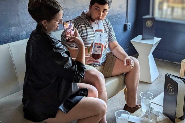 Two people sitting indoors, one taking a photo of the other drinking from a large beverage container.