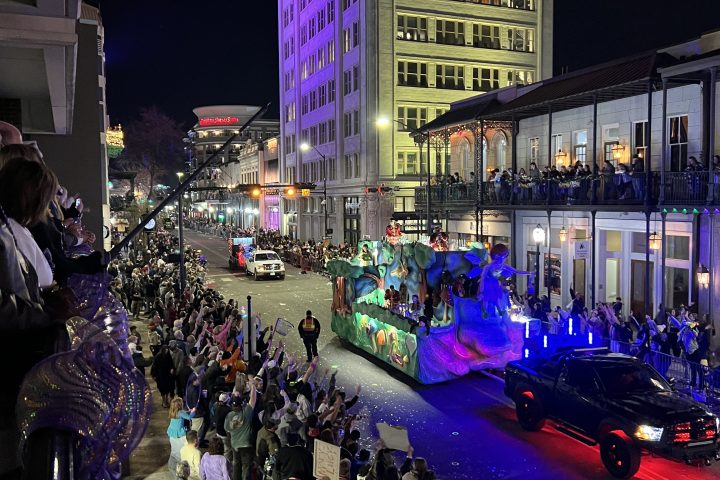 Night parade with colorful float, crowd cheering, tall lit building in background.