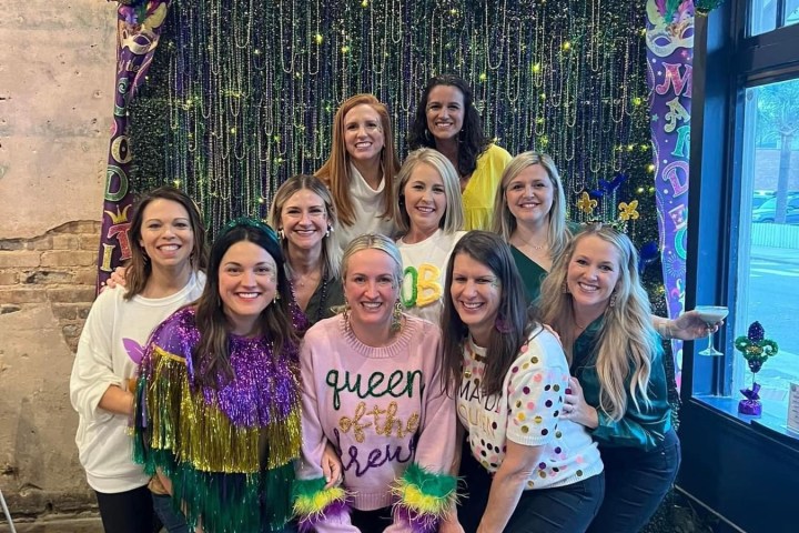 Group of women posing indoors with Mardi Gras-themed backdrop.