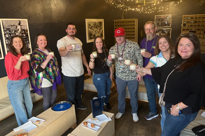 Group of nine people smiling and holding drinks in a cozy room with string lights.