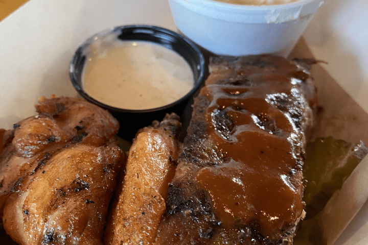 BBQ platter with ribs, chicken, mashed potatoes, and dipping sauce served in a paper tray.